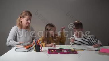 两名白人儿童和年轻女教师坐在桌子旁交谈的肖像。 教师教孩子们